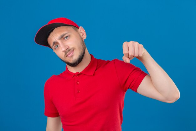 Young delivery man wearing red polo shirt and cap looking at camera smiling friendly gesturing fist bump as if greeting approving or as sign of respect over isolated blue background