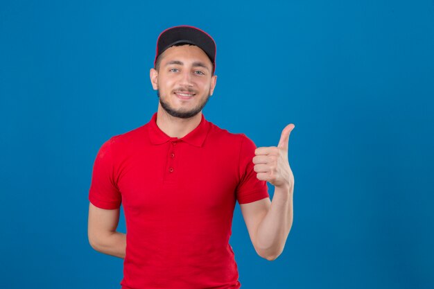 Young delivery man wearing red polo shirt and cap looking at camera with friendly smile showing thumb up over isolated blue background