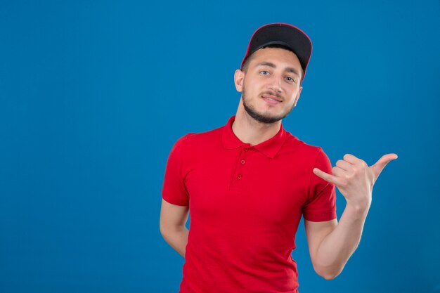 Young delivery man wearing red polo shirt and cap making call me gesture looking confident over isolated blue background
