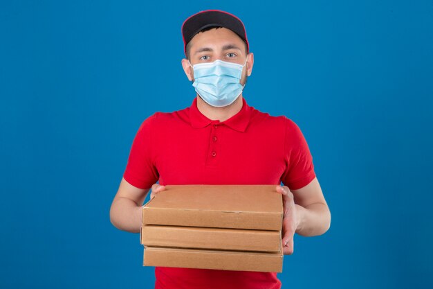 Young delivery man wearing red polo shirt and cap in protective medical mask standing with stack of pizza boxes looking at camera with serious face over isolated blue background