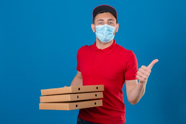 Young delivery man wearing red polo shirt and cap in protective medical mask standing with stack of pizza boxes showing thumb up over isolated blue background