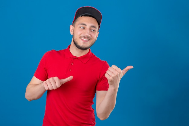 Young delivery man wearing red polo shirt and cap smiling with happy face looking and pointing to the side with thumbs up over isolated blue background
