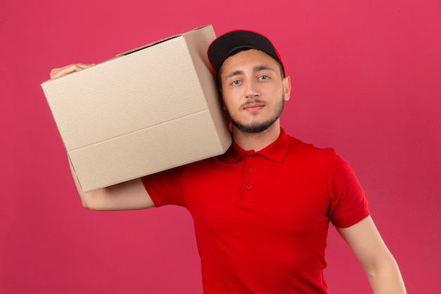 Free photo young delivery man wearing red polo shirt and cap standing with cardboard box on shoulder looking at camera with serious face over isolated pink background