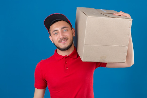 Free photo young delivery man wearing red polo shirt and cap standing with cardboard box on shoulder smiling looking at camera over isolated blue background