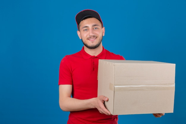 Young delivery man wearing red polo shirt and cap standing with cardboard smiling friendly looking at camera over isolated blue background