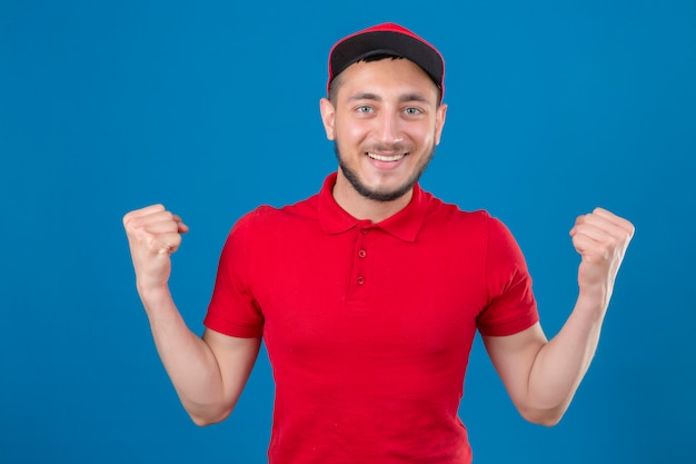 Young delivery man wearing red polo shirt and cap standing with fists raised looking confident winner concept over isolated blue background