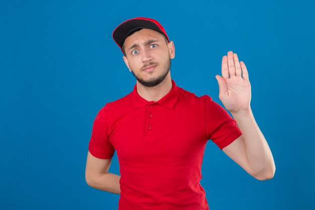 Young delivery man wearing red polo shirt and cap standing with open hand doing stop sign with serious and confident expression defense gesture over isolated blue background