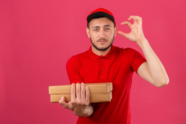 Young delivery man wearing red polo shirt and cap standing with pizza boxes looking at camera showing size sign with fingers over isolated pink background