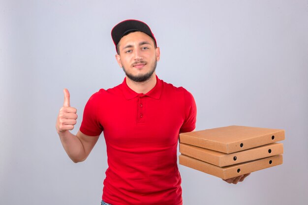 Молодой курьер в красной рубашке поло и кепке, стоящий со стопкой коробок для пиццы, выглядит уверенно, показывая большой палец вверх на изолированном фоне