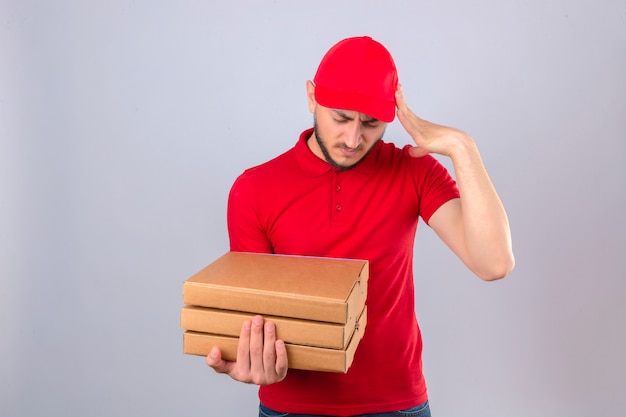 Young delivery man wearing red polo shirt and cap standing with stack of pizza boxes looking overworked touching head suffering from headache over isolated white background