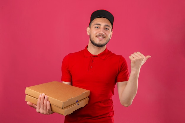 Молодой курьер в красной рубашке поло и кепке, стоящий с стопкой коробок для пиццы, указывая и показывая большим пальцем в сторону со счастливым лицом, улыбающимся на изолированном розовом фоне