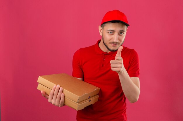 Young delivery man wearing red polo shirt and cap standing with stack of pizza boxes showing thumb up smiling friendly over isolated pink background