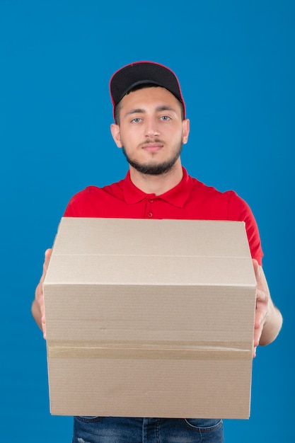 Молодой курьер в красной рубашке поло и кепке протягивает стопку коробки для пиццы с серьезным лицом на изолированном синем фоне