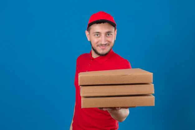 Young delivery man wearing red polo shirt and cap stretching out a stack of pizza boxes with smile on face over isolated blue background