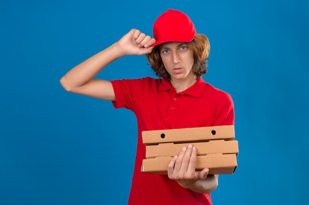 分離の青い背景に深刻な顔で彼の帽子に触れる挨拶ジェスチャーを作るピザの箱を保持している赤い制服を着ている若い配達人