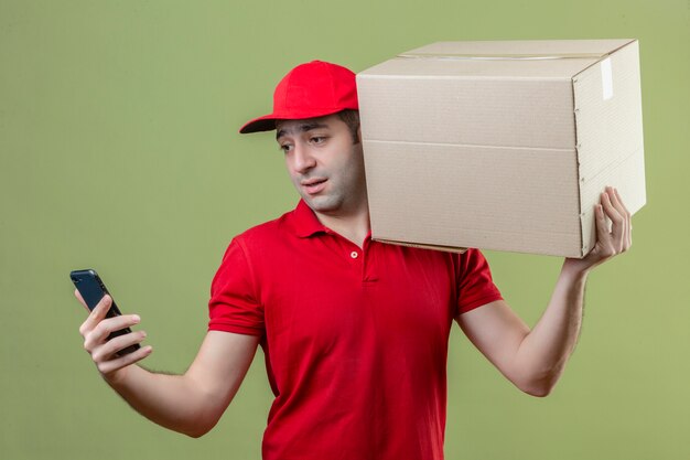 Young delivery man wearing red uniform standing with cardboard box on his shoulder looking at screen of his smartphone with sad expression on face over isolated green background