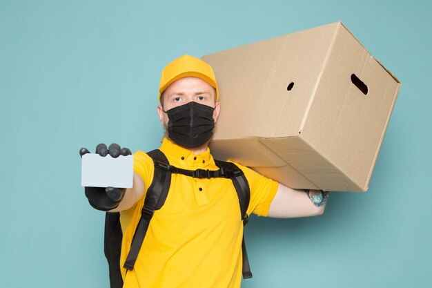 young delivery man in yellow polo yellow cap white jeans backpack and black sterile mask holding a box on blue