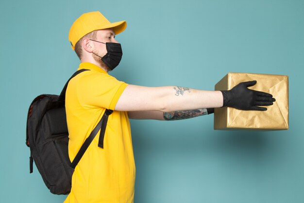 Молодой доставщик в желтой рубашке поло, желтой кепке, в белом джинсовом рюкзаке и черной стерильной маске с синей коробкой