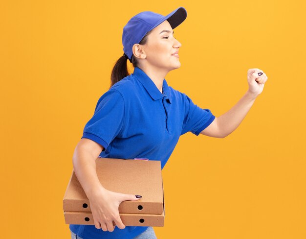 青い制服とキャップの若い配達の女性は、オレンジ色の壁を越えて顧客にピザの箱を配達するために急いで走っています