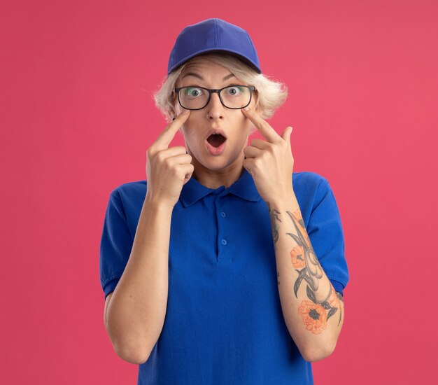 Young delivery woman in blue uniform and cap  being surprised pointing with index fingers at eyes over pink wall