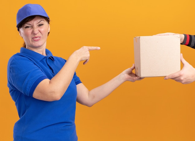 Young delivery woman in blue uniform and cap feeling confused while receiving box package pointing with index finger at box standing over orange wall