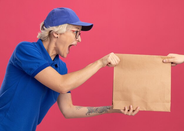 Young delivery woman in blue uniform and cap feeling excited while receiving paper package standing over pink wall