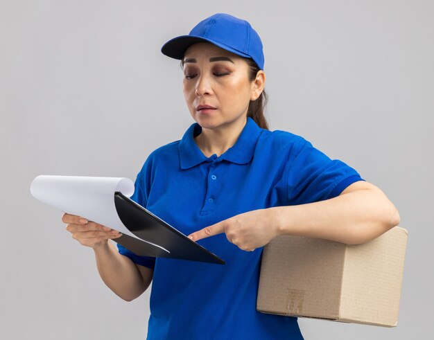 Young delivery woman in blue uniform and cap holding cardboard box and clipboard with blank pages looking at it with serious face standing over white wall