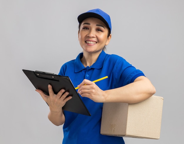 Молодая женщина-доставщик в синей форме и кепке держит картонную коробку и буфер обмена, пишет что-то уверенно улыбаясь, стоя над белой стеной