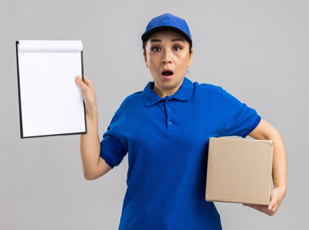 Young delivery woman in blue uniform and cap holding cardboard box showing clipboard   amazed and surprised  