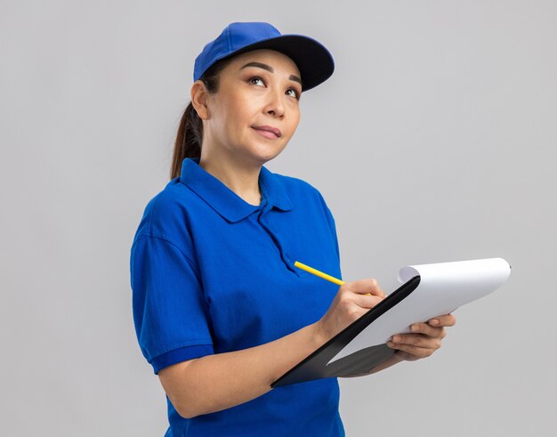 Молодая женщина-доставщик в синей форме и кепке держит в руках буфер обмена и ручку, озадаченно глядя на белую стену