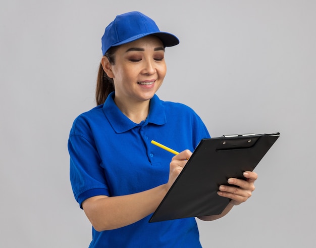 Giovane donna delle consegne in uniforme blu e berretto che tiene appunti e penna che sorride sicuro di scrivere qualcosa in piedi sul muro bianco