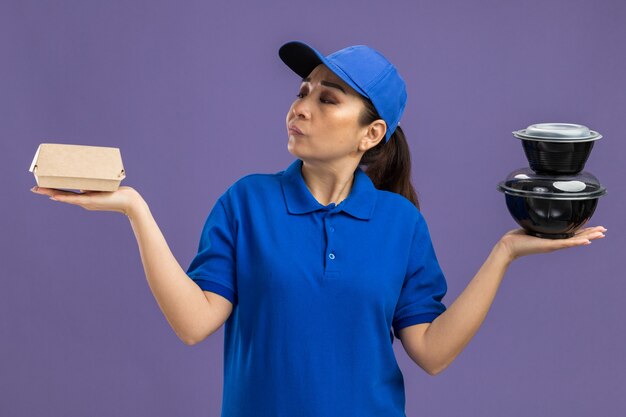 Young delivery woman in blue uniform and cap holding food packages looking confused having no answer standing over purple wall