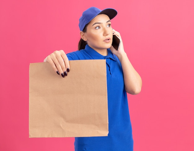 Молодая женщина-доставщик в синей форме и кепке держит бумажный пакет в замешательстве, разговаривая по мобильному телефону, стоя над розовой стеной