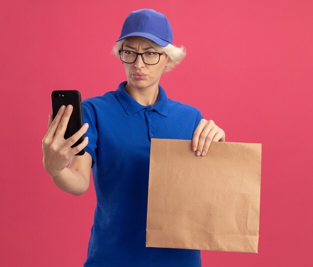 ピンクの壁に不満を持っている彼女のスマートフォンの画面を見ている青い制服とキャップ保持紙パッケージの若い配達女性