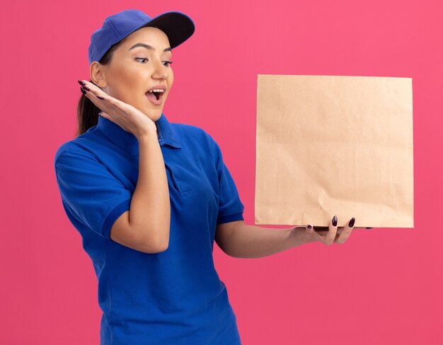 Молодая женщина-доставщик в синей форме и кепке держит бумажный пакет, глядя на него изумленно и удивленно, стоя над розовой стеной