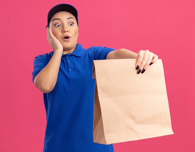 Молодая доставщица в синей униформе и кепке держит бумажный пакет, глядя на него, удивляясь и удивляясь, стоя у розовой стены