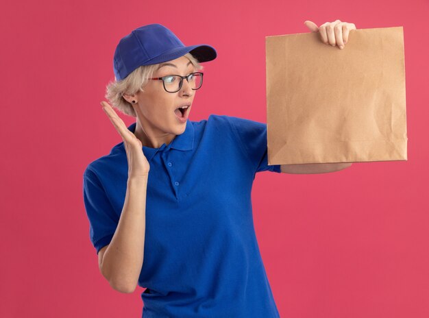 Молодая женщина-доставщик в синей форме и кепке держит бумажный пакет, глядя на него с удивлением и изумлением над розовой стеной