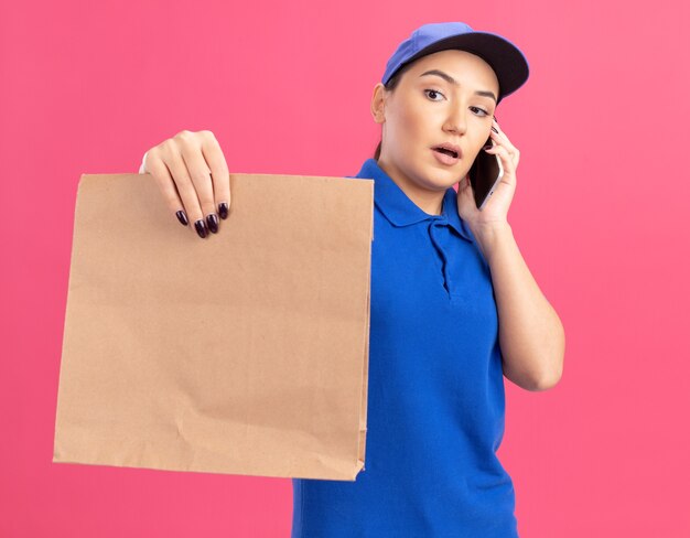 Giovane donna di consegna in uniforme blu e cappuccio che tiene il pacchetto di carta guardandolo con faccia seria mentre parla al telefono cellulare in piedi sopra il muro rosa