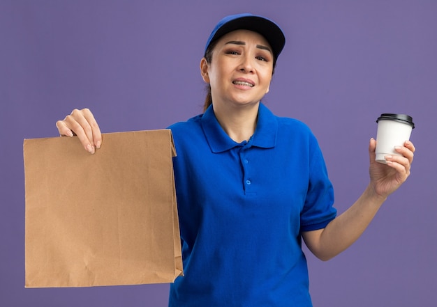 Young delivery woman in blue uniform and cap holding paper package and paper cup annoyed with sad expression standing over purple wall