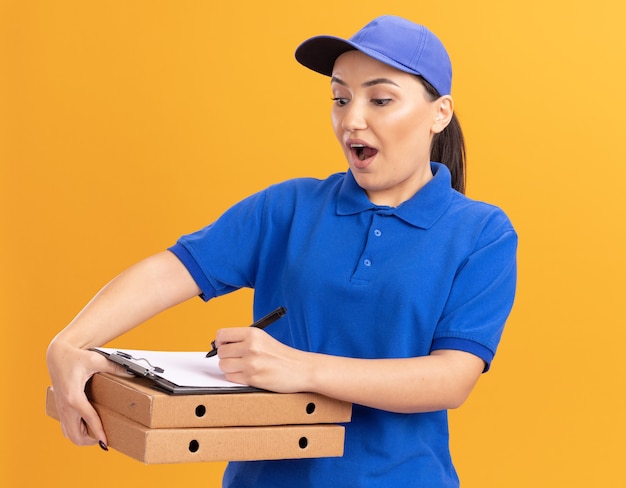 Young delivery woman in blue uniform and cap holding pizza boxes and clipboard with pencil writing happy and excited standing over orange wall