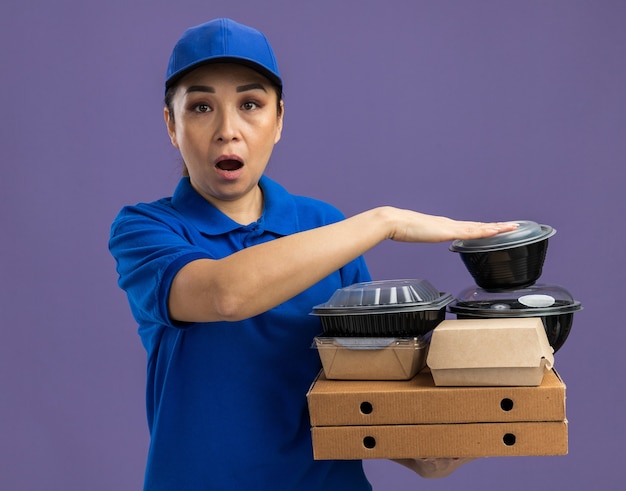 Молодая доставщица в синей форме и кепке, держащая коробки для пиццы и продуктовые пакеты, изумлена и удивлена, стоя у фиолетовой стены