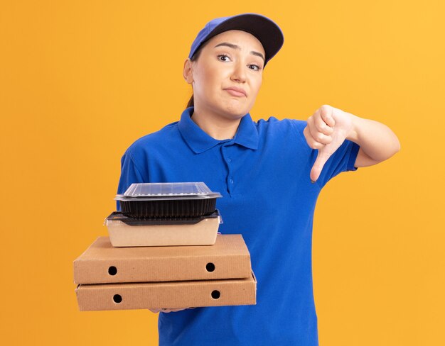 Молодая женщина-доставщик в синей форме и кепке держит коробки для пиццы и продуктовые пакеты, глядя вперед, смущенная и недовольная, показывая большие пальцы вниз, стоя над оранжевой стеной