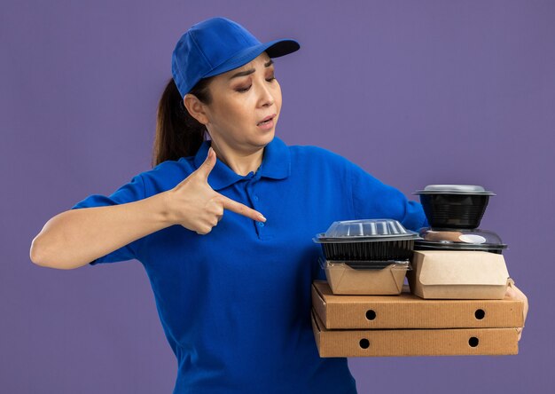 Young delivery woman in blue uniform and cap holding pizza boxes and food packages pointing with index finger at them standing over purple wall