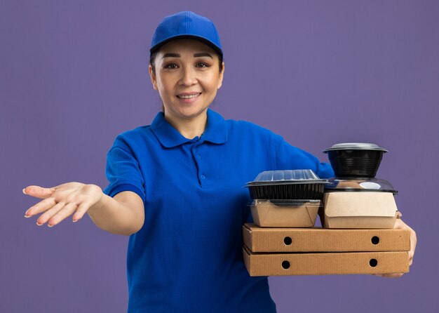 Young delivery woman in blue uniform and cap holding pizza boxes and food packages  with arm out smiling cheerfully standing over purple wall