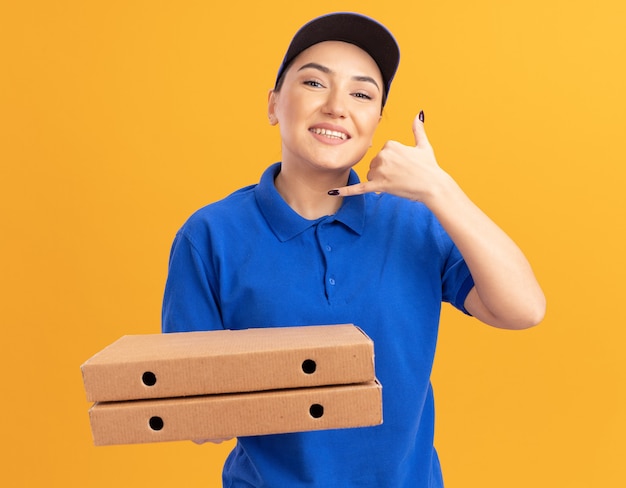 Young delivery woman in blue uniform and cap holding pizza boxes looking at front smiling cheerfully showing call me gesture with hand standing over orange wall