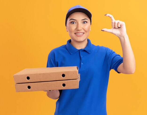 파란색 제복을 입은 젊은 배달 여자와 모자는 오렌지 벽 위에 유쾌하게 서있는 정면을보고 작은 크기의 제스처를 만드는 피자 상자를 들고