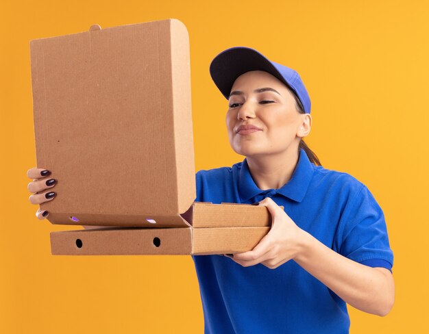 Молодая доставщица в синей форме и кепке держит коробки для пиццы, открывая коробку, вдыхая приятный аромат, стоя над оранжевой стеной