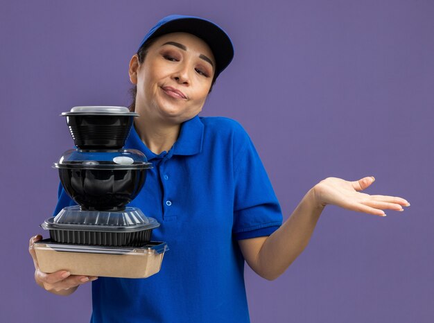 Молодая доставщица в синей форме и кепке держит стопку продуктовых пакетов, глядя на них в замешательстве, пожимая плечами, не имея ответа, стоя над фиолетовой стеной