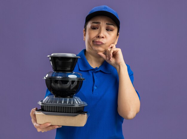 Young delivery woman in blue uniform and cap holding stack of food packages looking at them confused standing over purple wall