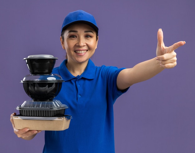 Young delivery woman in blue uniform and cap holding stack of food packages  smiling cheerfully pointing with index finger  standing over purple wall